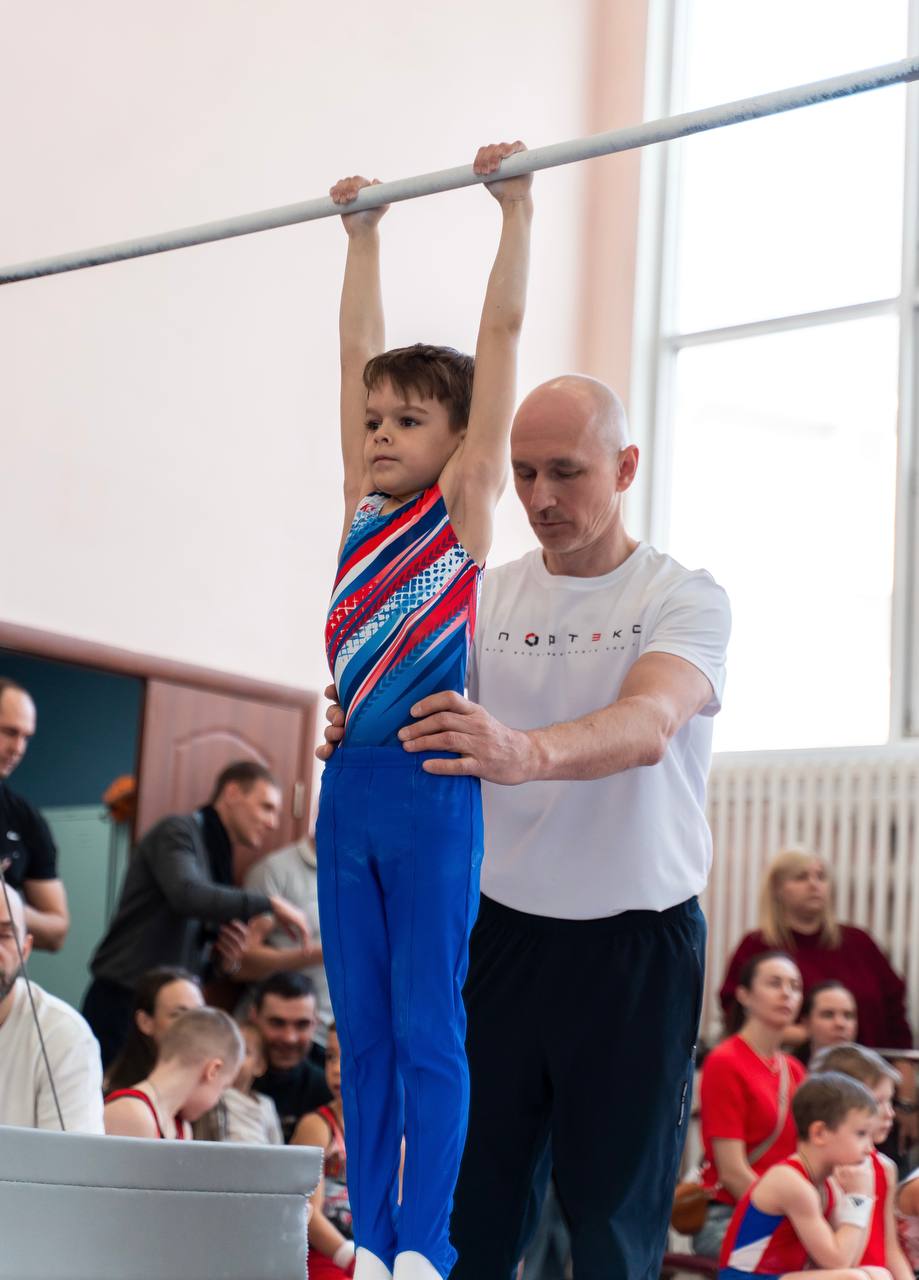 Тренер помогает юному гимнасту в синей форме удерживать положение на перекладине